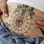 Piercing Bananabell plateado en un ombligo con tatuaje de telaraña y jeans.