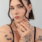 Donna con capelli corti, collana d'argento e diversi anelli, braccia tatuate, sfondo neutro
