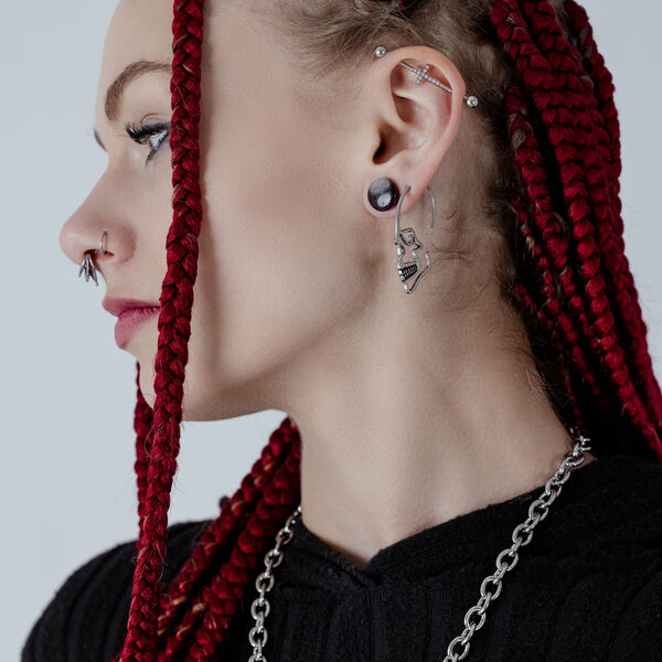 Pendiente de calavera plateado de acero quirúrgico en la oreja de una persona con trenzas rojas