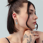 Donna con capelli corti e scuri indossa creole argento con design a cuore e tatuaggi su sfondo grigio chiaro.