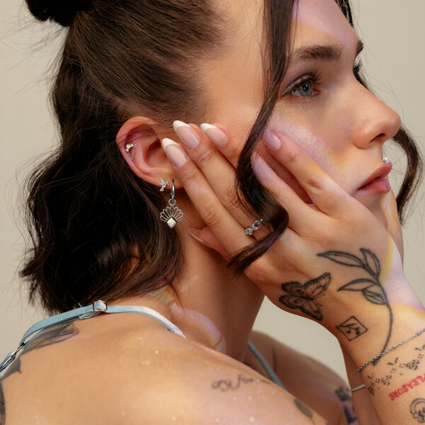 Giovane donna con luce sfumata sul viso, indossa un orecchino d'argento con ciondolo a farfalla e ha tatuaggi sulle mani.