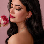 Mujer con cabello largo y oscuro sostiene una rosa rosa y lleva un pendiente de plata con cristal rojo sobre fondo rosa