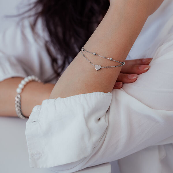 Pulsera plateada de dos hileras con colgante de corazón en el brazo de una persona con ropa blanca
