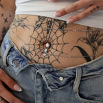 Banana Bell in argento con cristalli, indossato su un ventre tatuato con jeans.
