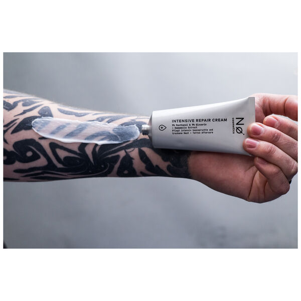 Tubo gris-blanco de la Crema Reparadora Intensiva de No Cosmetics, sostenido por una mano tatuada