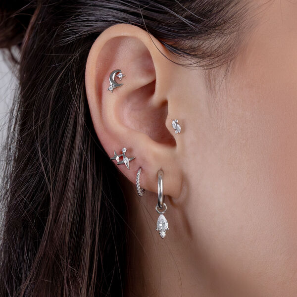 Nahaufnahme eines Ohrs mit mehreren silbernen Piercings und kristallklaren Steinchen, auf braunem Haarhintergrund.