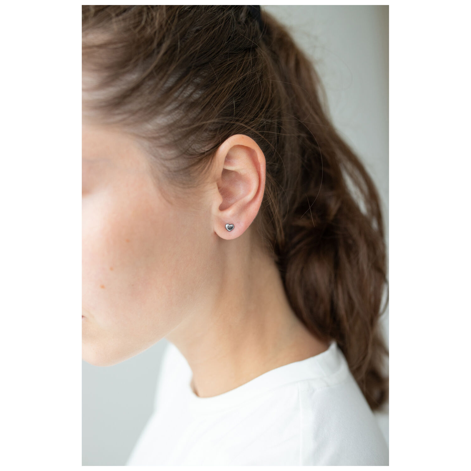 Herz Ohrstecker aus Edelstahl in Silber Silberne Herz Ohrstecker am Ohr einer Frau mit braunen Haaren, weißer Hintergrund