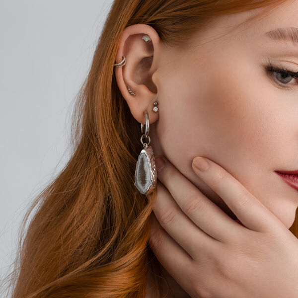 Primer plano de una oreja con varios piercings de plata y un colgante de geoda.